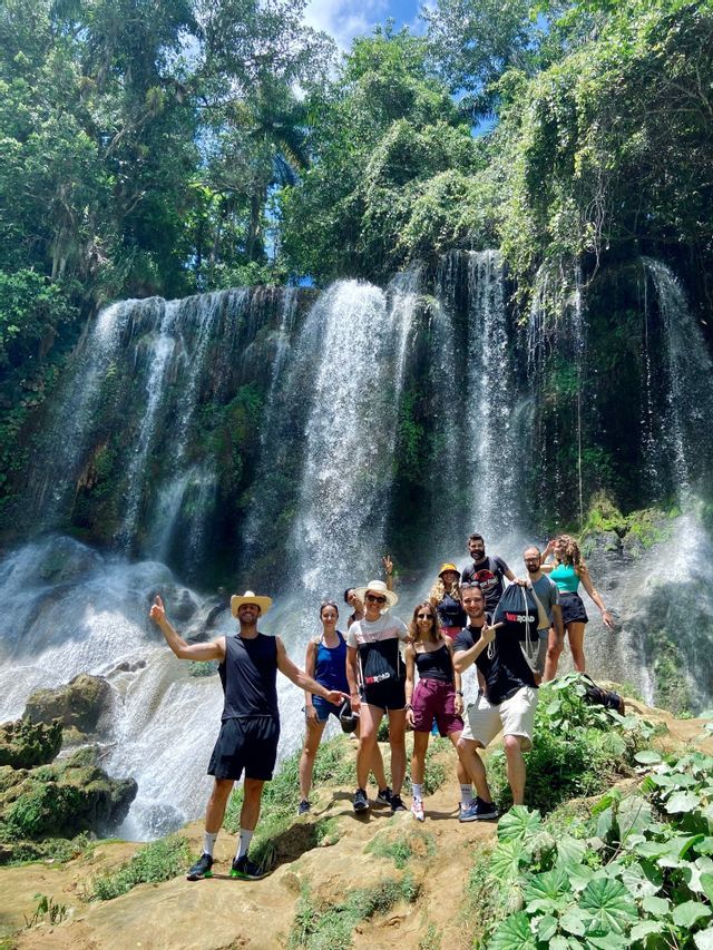 Eine WeRoad-Gruppe posiert für ein Foto auf einem Felsvorsprung vor einem großen, kaskadierenden Wasserfall in einem üppigen Wald.