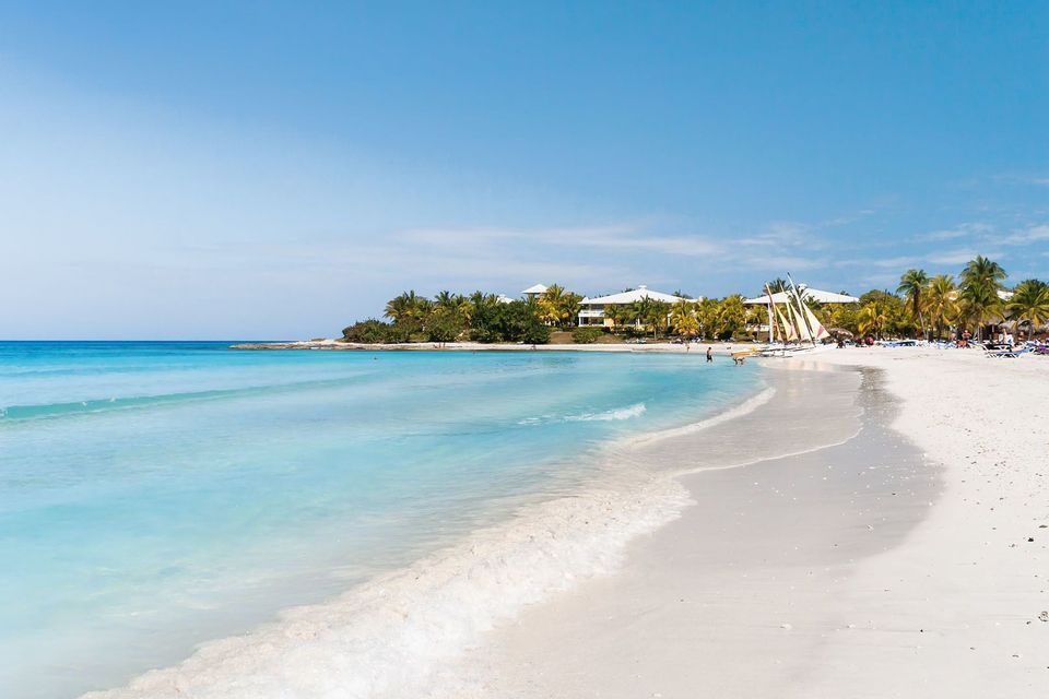 Una spiaggia di sabbia bianca con acque turchesi che lambiscono la riva. Sullo sfondo si ergono palme, barche a vela ed edifici di resort.