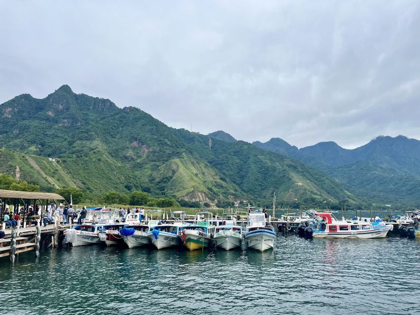 Una flota de pequeños barcos está atracada en un muelle de madera en un lago, con grandes montañas verdes elevándose al fondo bajo un cielo nublado.