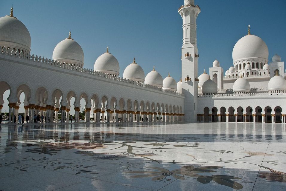 Eine große weiße Moschee mit mehreren Kuppeln und einem Minarett, die sich in einem polierten Innenhof mit floralen Mosaiken spiegelt.