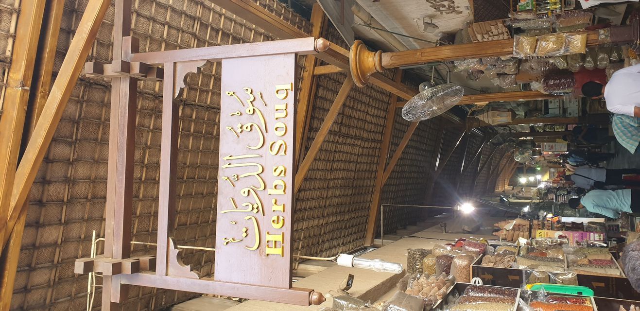 Ein Holzschild für einen 'Kräuter-Souk' hängt am Reetdach eines geschäftigen, überdachten Marktes voller Gewürzstände.