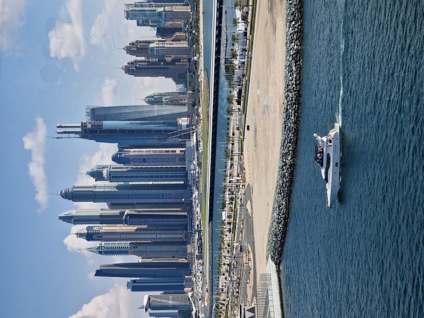 Eine Luftaufnahme zeigt eine weiße Yacht, die in blauem Wasser neben einer modernen Stadtkulisse unter einem teilweise bewölkten Himmel segelt.