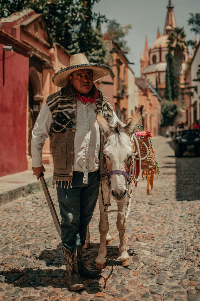 Ein Mann mit Sombrero und traditioneller Kleidung geht neben einem weißen Esel auf einer Kopfsteinpflasterstraße.
