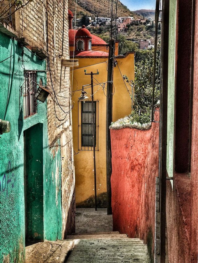 Eine schmale Gasse mit Steinstufen führt zwischen farbenfrohen türkisfarbenen, gelben und roten Gebäuden hinunter, mit Blick auf eine Stadt am Hang.