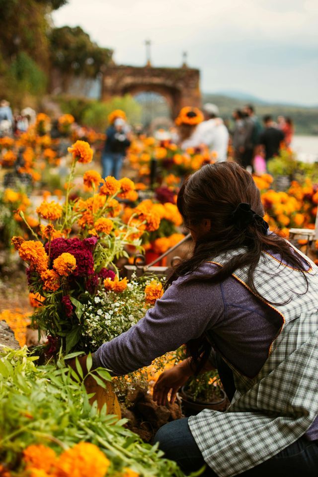 Eine Person mit einer karierten Schürze kniet im Freien, um einen großen Strauß aus orangefarbenen Ringelblumen und roten Blumen zu arrangieren.