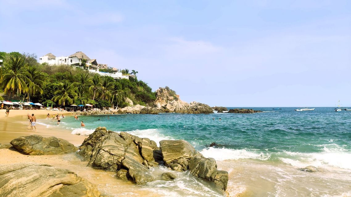 Ein Blick auf eine tropische Strandbucht, wo Menschen in türkisfarbenem Wasser neben einem Ufer mit Felsen und Palmen schwimmen.