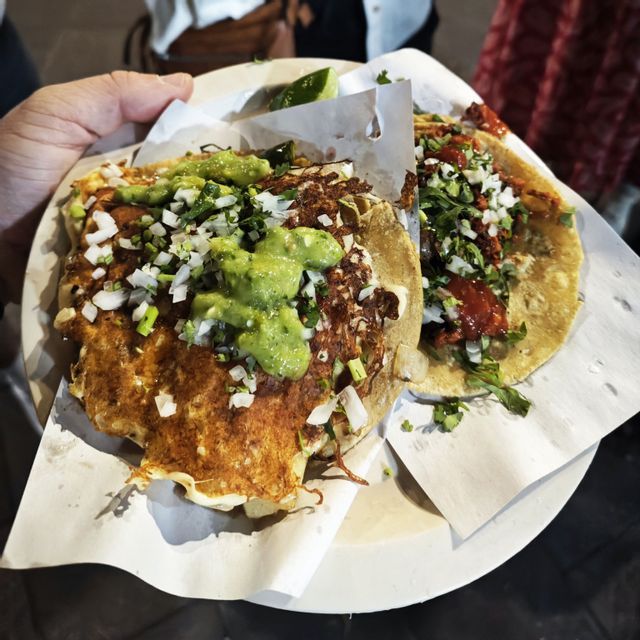 Eine Hand hält einen Pappteller mit zwei Tacos, belegt mit knusprigem Käse, gehackten Zwiebeln, Koriander und grüner Salsa.
