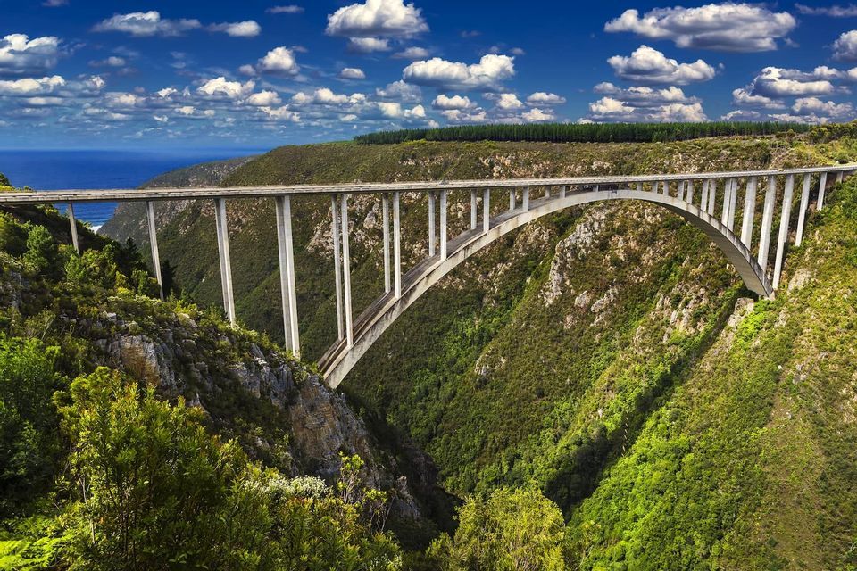 Eine lange Betonbogenbrücke überspannt eine tiefe, von üppig grünem Laub bedeckte Schlucht, mit dem Ozean in der Ferne unter einem blauen, bewölkten Himmel.