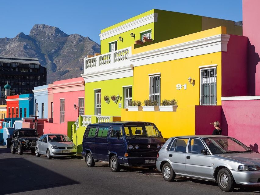 Des voitures sont garées dans une rue bordée de bâtiments vifs et colorés, devant une grande montagne sous un ciel bleu clair.