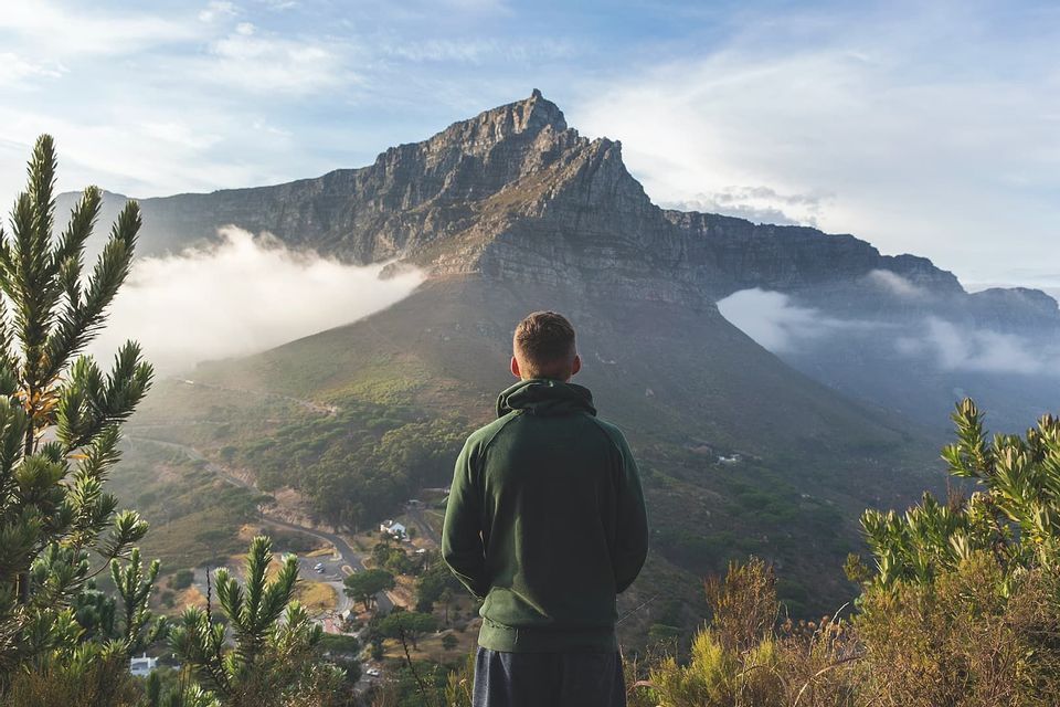 A person in a green hoodie stands on a viewpoint, gazing at a large, rocky mountain peak partially covered by clouds.