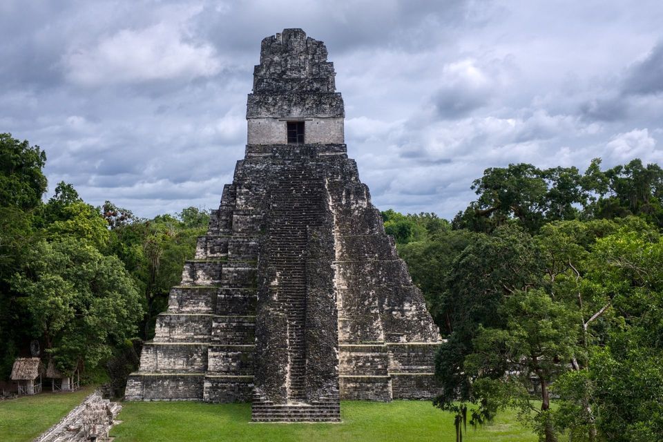 Una gran pirámide escalonada de piedra se alza desde un claro verde en una densa jungla bajo un cielo nublado.