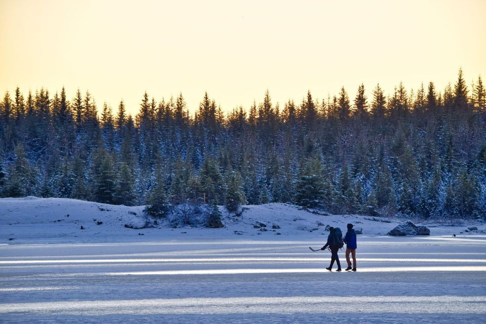 Zwei Personen mit Rucksäcken, eine davon mit einem Hockeyschläger, gehen über einen zugefrorenen See vor einem schneebedeckten Kiefernwald.