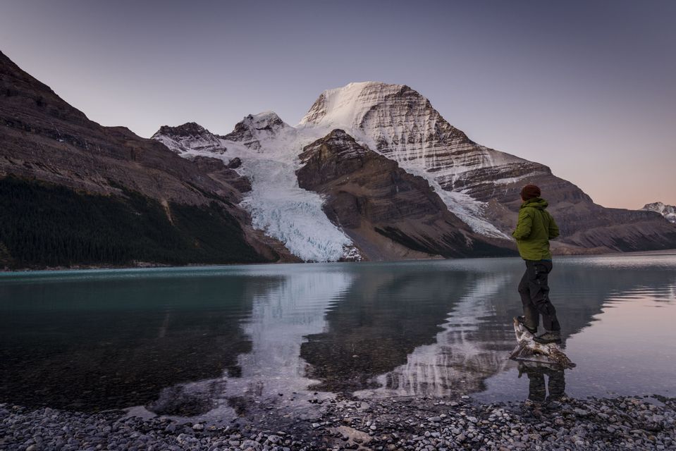 Eine Person steht an einem felsigen Ufer und blickt über einen ruhigen See auf einen schneebedeckten Berg mit einem großen Gletscher.
