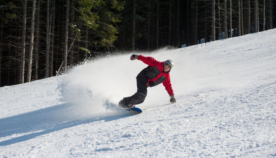 Ein Snowboarder in roter Jacke fährt eine verschneite Piste hinunter, wirbelt Pulverschnee auf, mit einem Kiefernwald im Hintergrund.