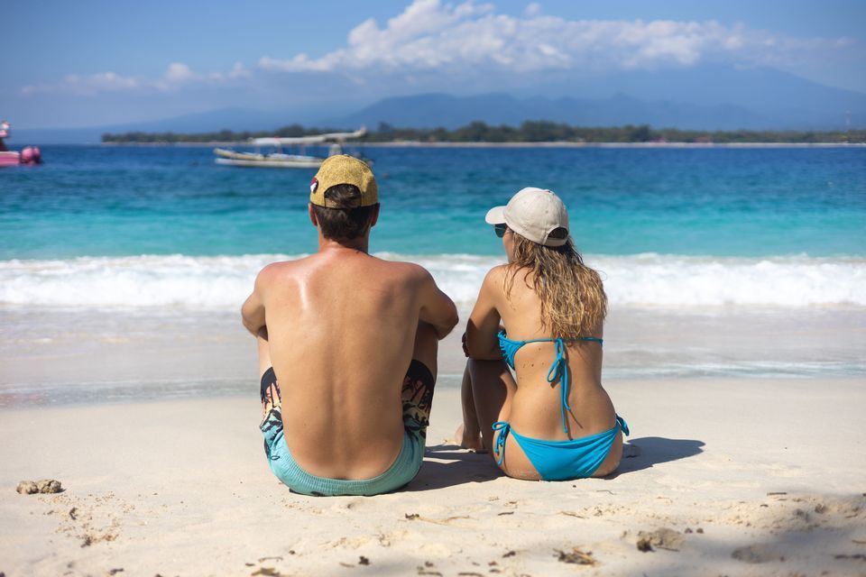 Un hombre y una mujer sentados en una playa de arena blanca, contemplando el océano turquesa y las montañas distantes en un día soleado.