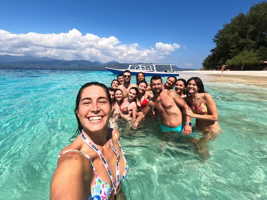 Una mujer se toma una selfie con su grupo WeRoad mientras sonríen juntos en las claras aguas turquesas de una playa tropical.