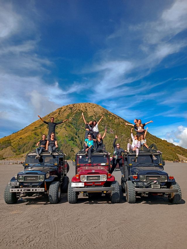 Eine WeRoad-Reisegruppe posiert auf drei Geländewagen vor einem großen, grasbewachsenen Berg unter einem teilweise bewölkten Himmel.