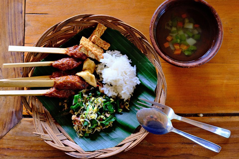 Un plato de brochetas de satay, tempeh, arroz y ensalada en un plato tejido, con un acompañamiento de sopa de verduras.