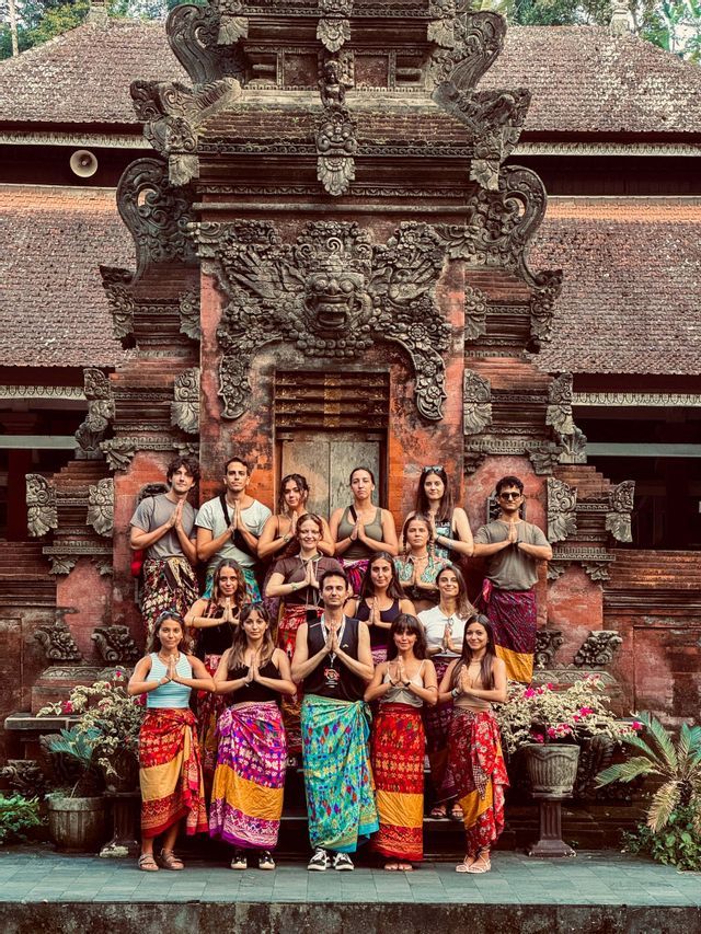Un grupo de WeRoad con pareos coloridos posa con las manos en gesto de oración frente a un templo de piedra ornamentado.