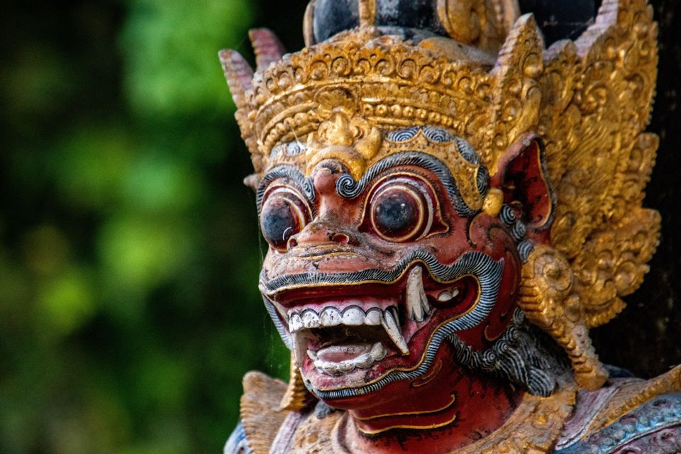 Primer plano de una estatua tradicional de guardián asiático con un rostro rojo, colmillos al descubierto y un tocado dorado ornamentado.