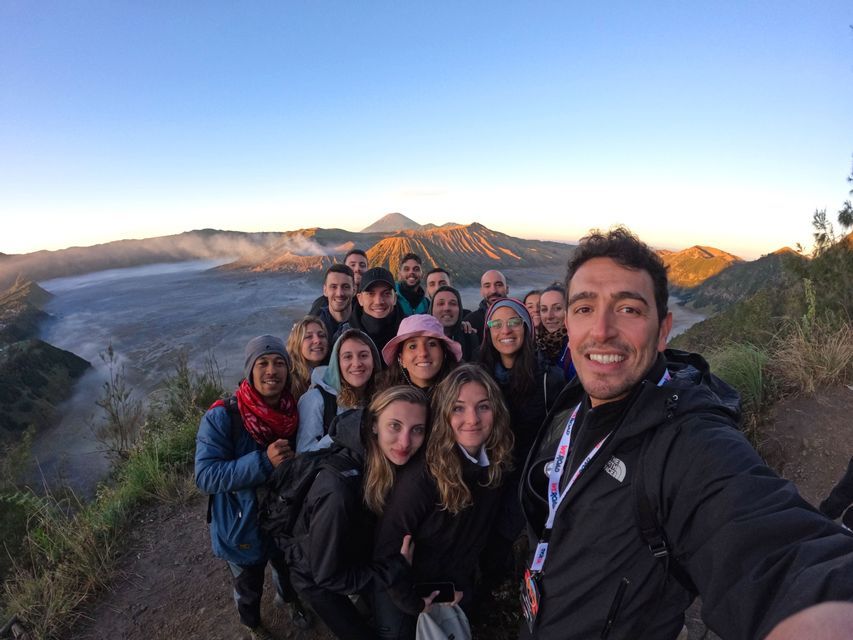 Un grupo de viaje de WeRoad se toma una selfie en un mirador, con un paisaje de caldera volcánica al fondo al amanecer.