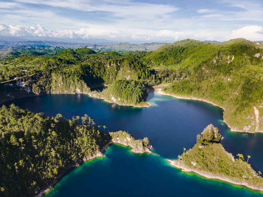 An aerial view of a winding system of deep blue lakes surrounded by lush, forested green hills under a cloudy sky.