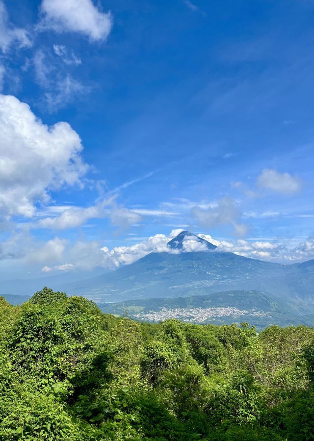 Un gran volcán se alza sobre un pueblo en el valle, visto sobre un exuberante bosque verde bajo un cielo azul parcialmente nublado.