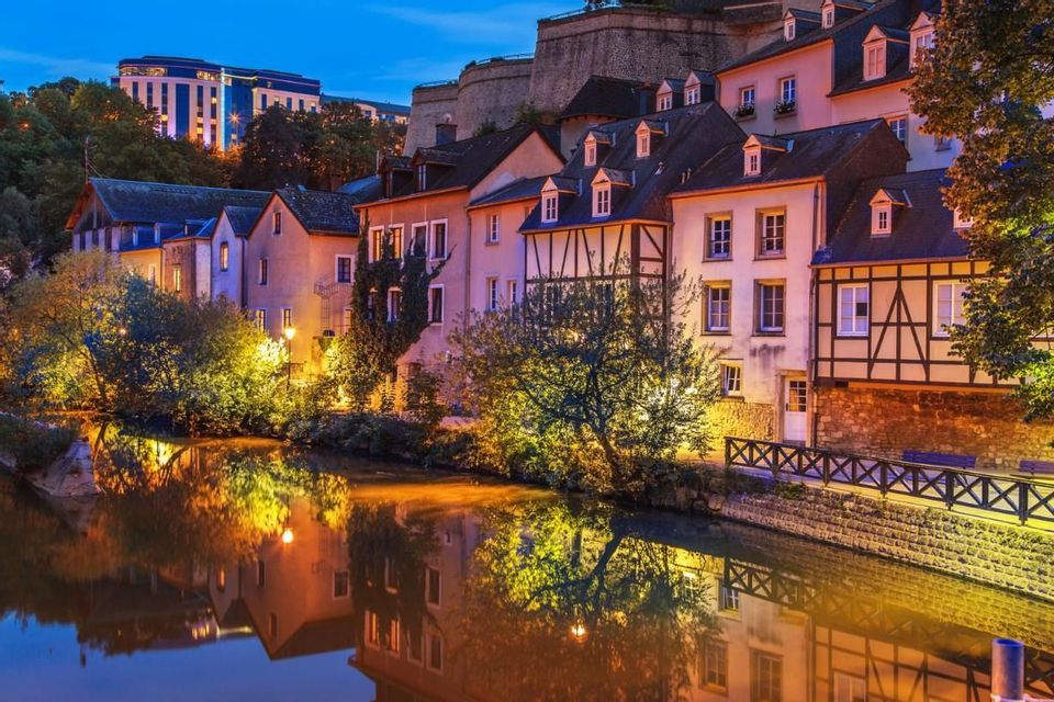 Edifici storici illuminati e alberi che costeggiano un fiume, con le luci riflesse sull'acqua calma al tramonto.