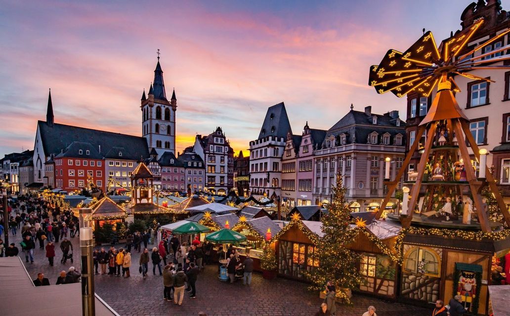 Un vivace mercatino di Natale su una piazza lastricata al tramonto, con bancarelle illuminate, una grande chiesa e una tradizionale piramide di legno.