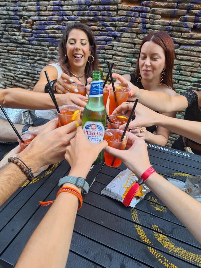 A WeRoad group trip toasting with orange cocktails and a beer bottle over a black table outdoors.