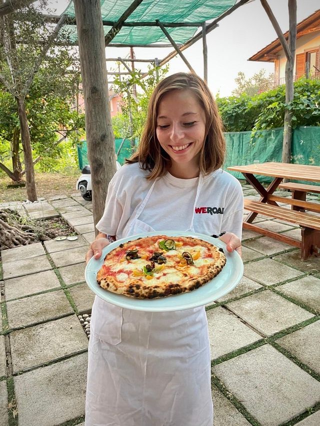 A smiling woman wearing a white apron and a WeRoad t-shirt holds a plate with a pizza on it while standing on an outdoor patio.