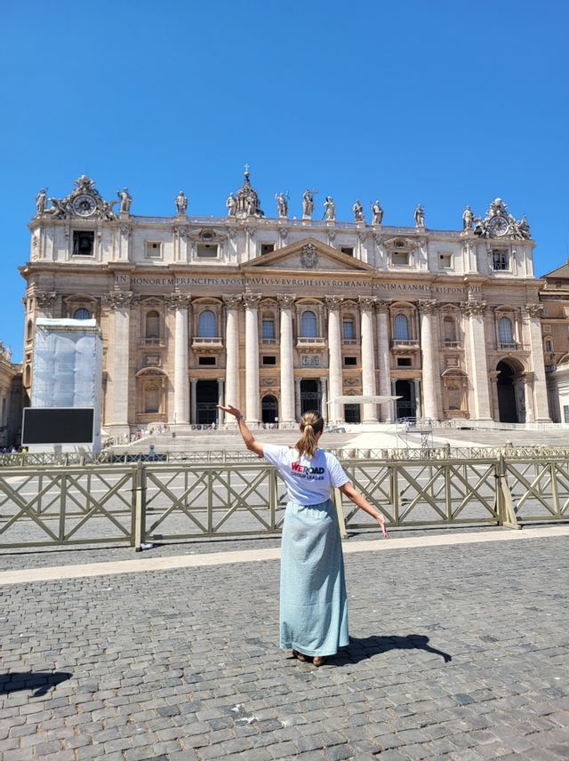 A woman wearing a WeRoad Group Leader t-shirt stands with her back to the camera, arms outstretched towards a grand basilica.
