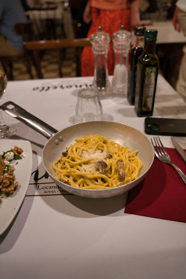 A pan of spaghetti carbonara topped with grated cheese sits on a set restaurant table with a red napkin.