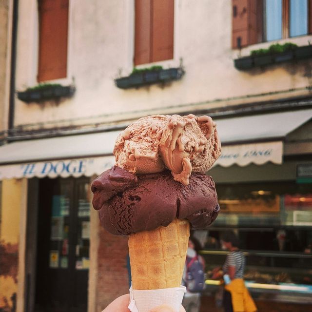 A close-up of a hand holding a waffle cone with a scoop of dark chocolate gelato and a scoop of milk chocolate gelato in front of a storefront.