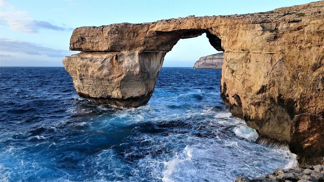 Une vaste arche de pierre naturelle surplombe une mer bleue agitée, avec des vagues qui se brisent au pied des falaises.