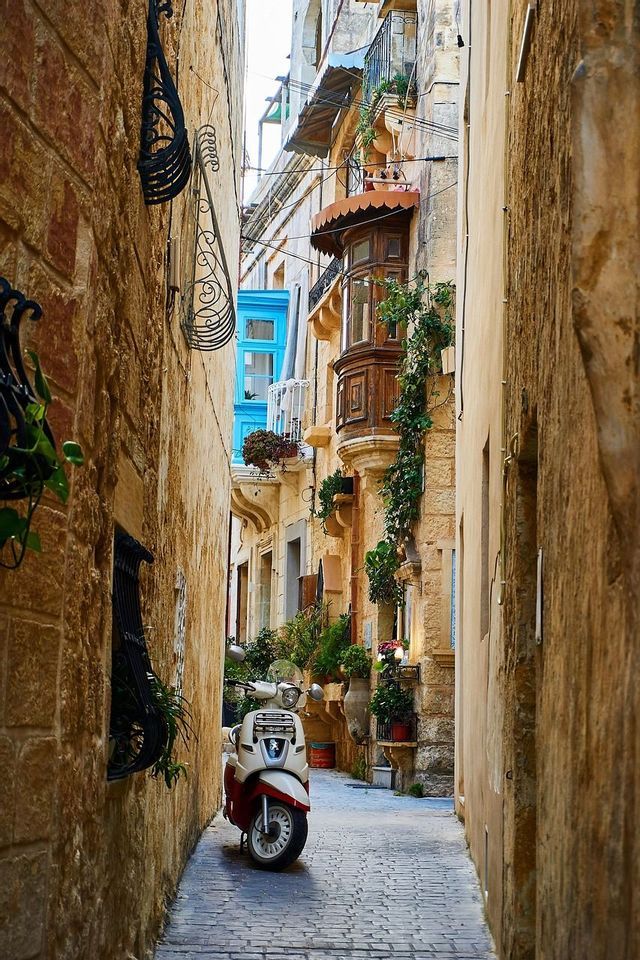 Un scooter rouge et blanc garé sur un chemin pavé dans une ruelle étroite entre de hauts bâtiments en pierre.