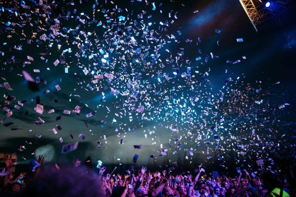 Des confettis pleuvent sur une foule immense, les mains levées, lors d'un concert nocturne sous des lumières de scène bleues.