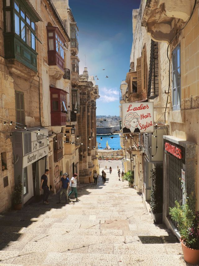 Une vue plongeante sur une rue pavée étroite et escarpée, bordée de vieux bâtiments avec balcons, menant à un port bleu et ensoleillé.