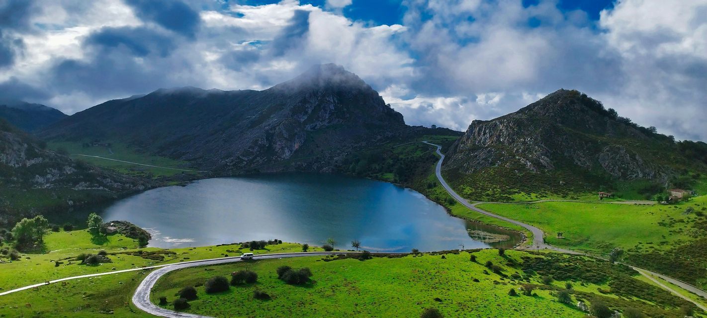 Una vista aerea di una strada tortuosa lungo un lago calmo, incastonata tra verdi colline e montagne sotto un cielo nuvoloso.