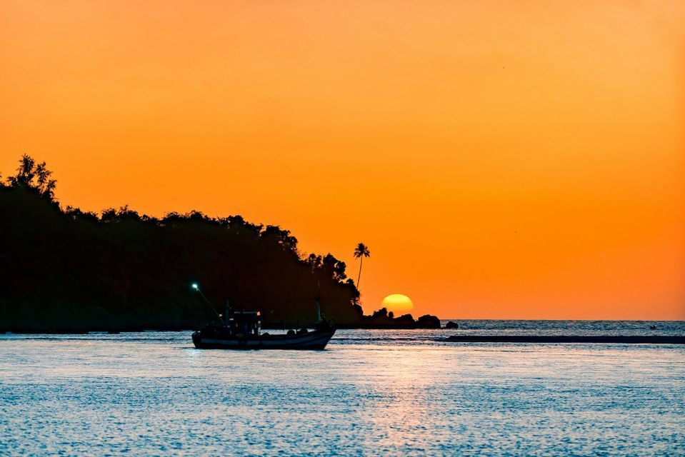 Una barca da pesca galleggia sull'acqua al tramonto, con una costa e una palma in silhouette sullo sfondo sotto un cielo arancione.