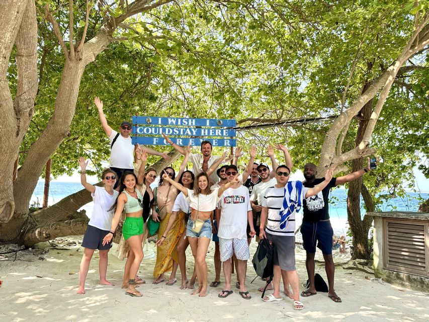 Un groupe WeRoad pose en souriant sur une plage de sable sous un grand arbre, avec un panneau bleu et l'océan en arrière-plan.