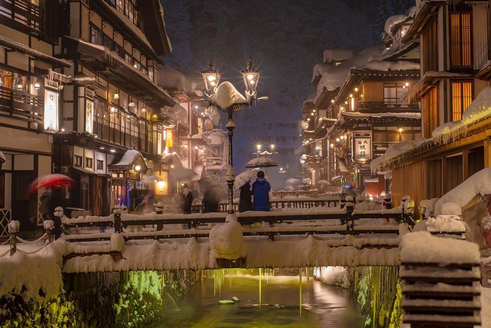 Un villaggio innevato di notte, con edifici tradizionali, un ponte su un canale e lanterne illuminate.