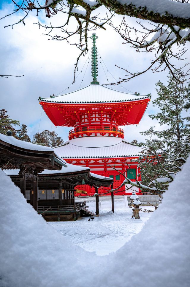 Eine rote japanische Pagode mit schneebedeckten Dächern und verschneiter Umgebung, gesehen durch einen natürlichen Rahmen aus schneebedeckten Ästen.