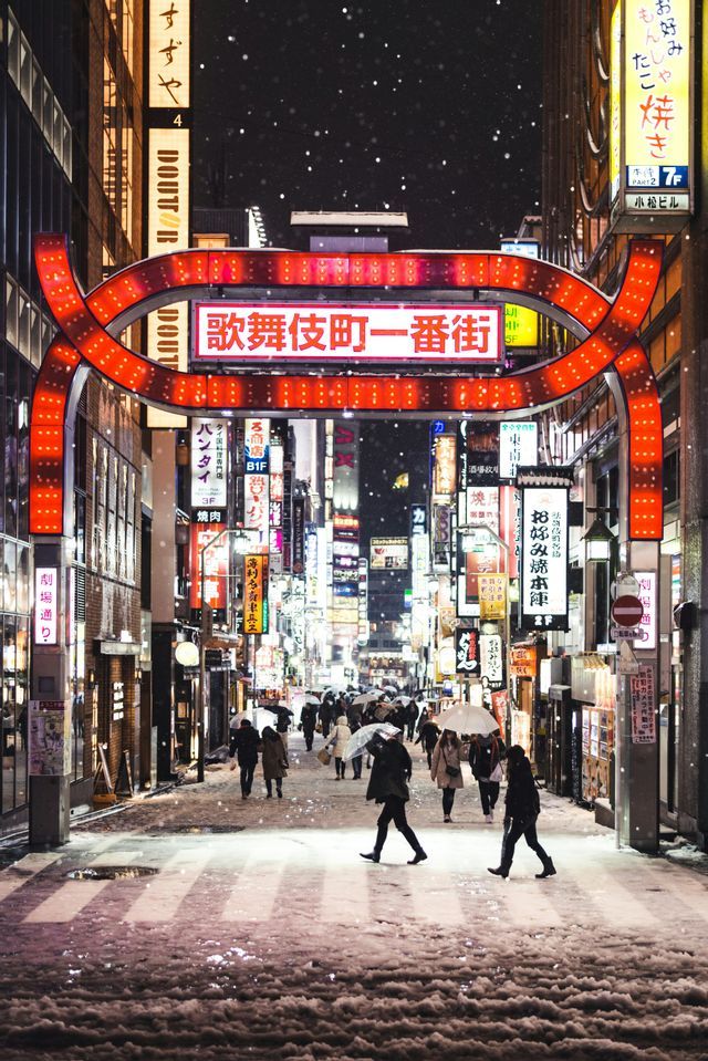Menschen gehen nachts auf einer verschneiten Stadtstraße unter einem großen roten Torbogen und leuchtenden Schildern mit asiatischen Schriftzeichen.