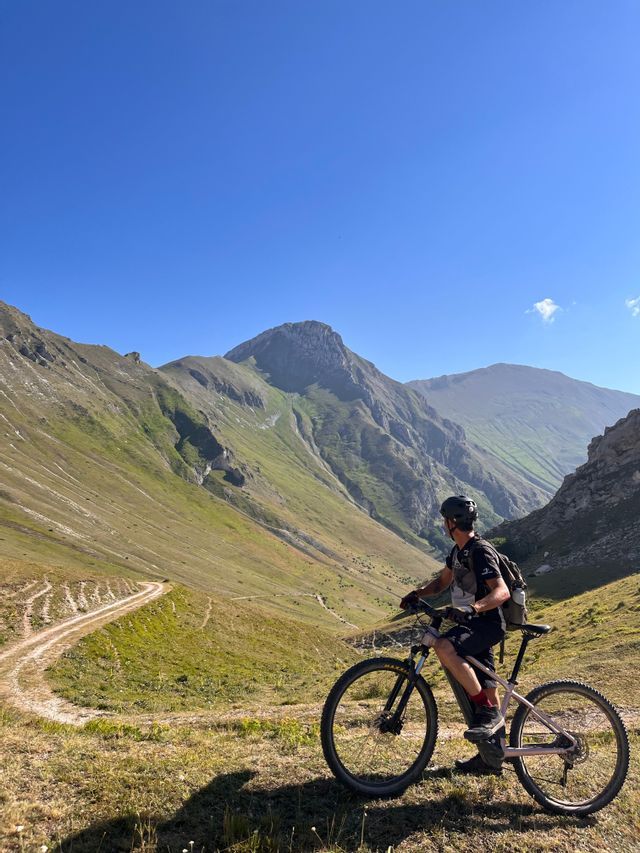 Un ciclista con casco e zaino in sosta sulla mountain bike, con vista su una vasta valle montana verde e un sentiero sterrato.