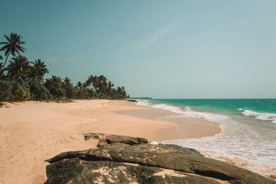 Una vista da dietro grandi rocce di una spiaggia sabbiosa costeggiata da palme, accanto a un oceano turchese sotto un cielo sereno.