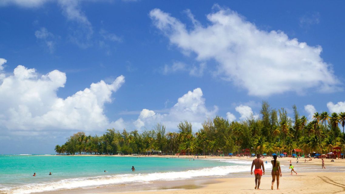 Due persone camminano lungo la riva di una spiaggia tropicale con acqua turchese, palme e un cielo azzurro e nuvoloso.