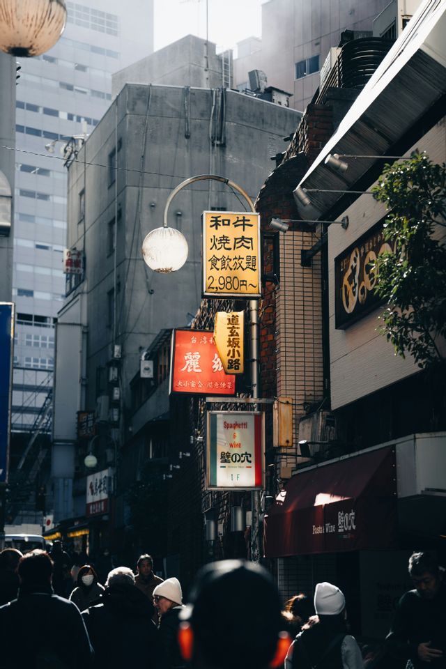 Eine Menschenmenge geht eine schmale Stadtstraße entlang, die von Gebäuden und vertikalen Schildern mit japanischen Schriftzeichen gesäumt ist.