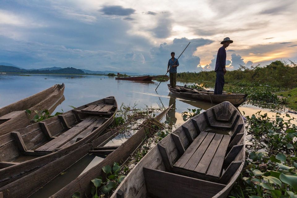 Dos hombres en un bote de madera en un lago tranquilo, rodeados de otras embarcaciones y plantas acuáticas, durante un atardecer nublado.