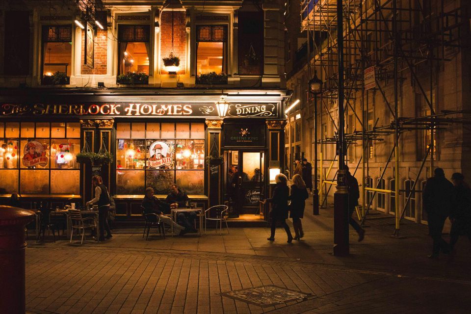 The exterior of 'The Sherlock Holmes' pub at night, with people sitting at outdoor tables and walking on the sidewalk.